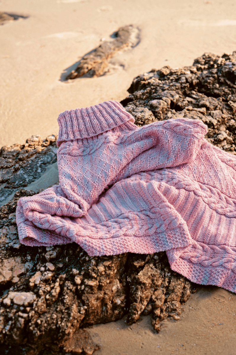 The Timeless Knits sweater by Malabrigo Yarn, made from hand-dyed Malabrigo yarn and featuring textured patterns, is draped over rugged rocks on a sandy beach with gentle sunlight casting soft shadows.