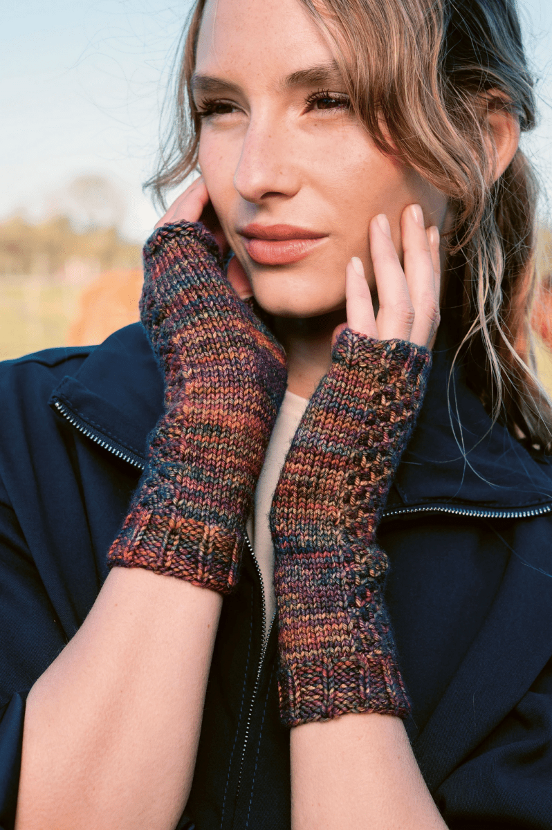 A woman in a dark jacket poses outdoors, showing off her loose waves and wearing Timeless Knits, Malabrigo fingerless gloves by Malabrigo Yarn—vibrant knit accessories crafted from hand-dyed yarn.