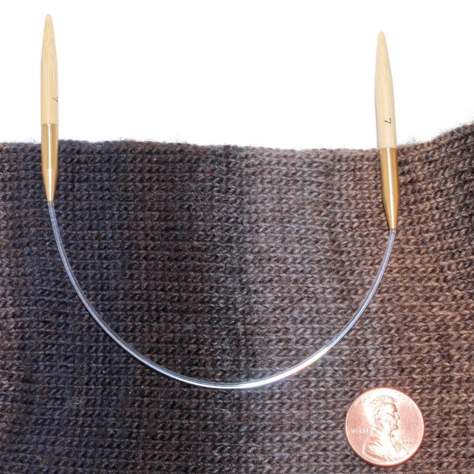 The Accessories Unlimited Circular Bamboo Knitting Needles by SeeKnit, with wooden tips and a flexible cord, are shown on dark variegated knitted fabric next to a U.S. penny for size comparison.