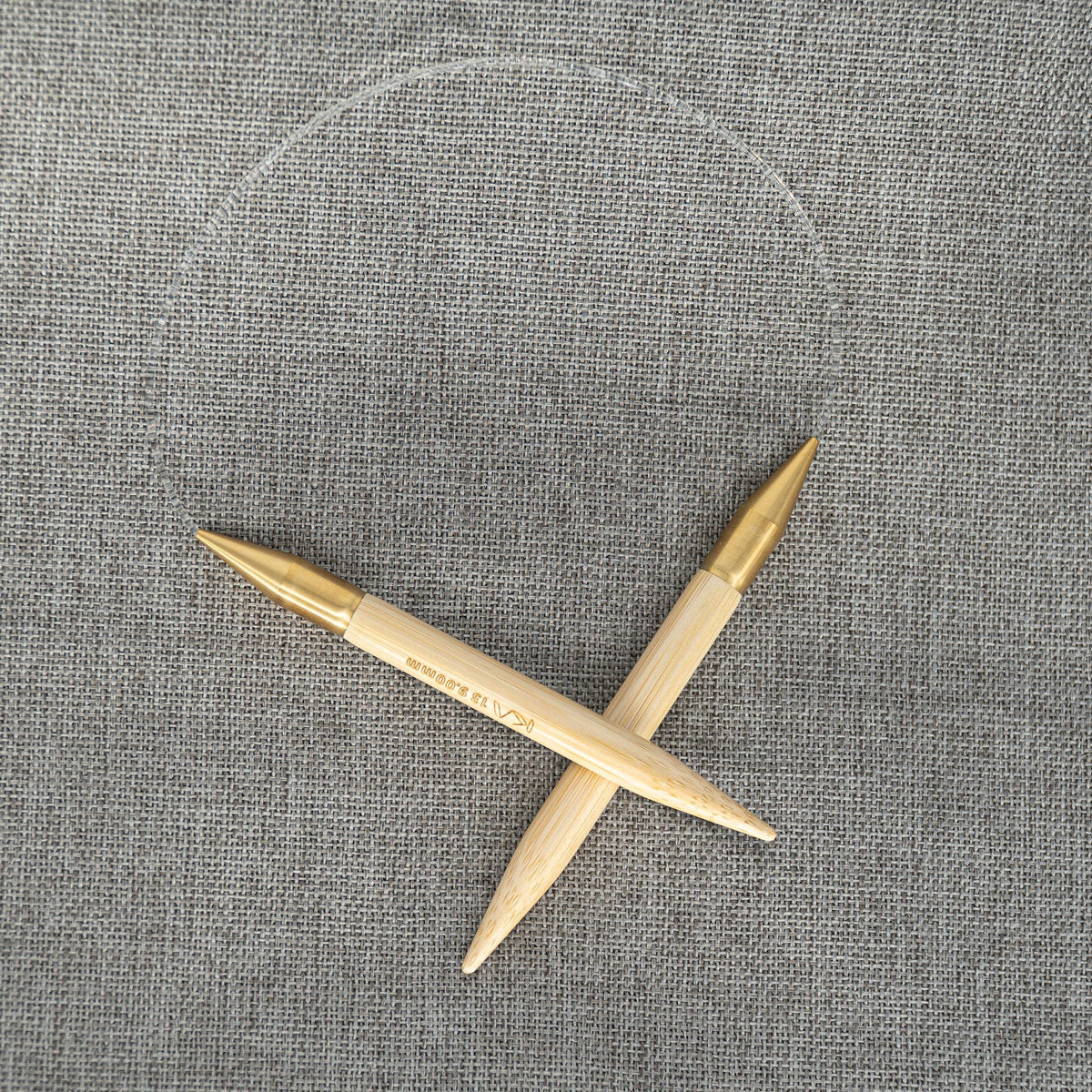 Two Accessories Unlimited Circular Bamboo Knitting Needles (SeeKnit), with gold tips and a clear cable with a flexible rotating joint, are crossed and displayed on textured grey fabric.
