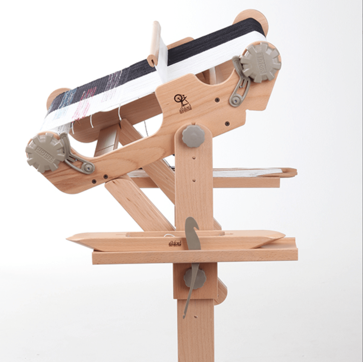 A wooden tabletop loom with yarn threads partially woven and a shuttle on a lower shelf is shown. It pairs well with the Ashford Rigid Heddle Loom Stand Accessories Tray from Ashford Handicrafts Limited for organizing weaving tools. Plain white background.