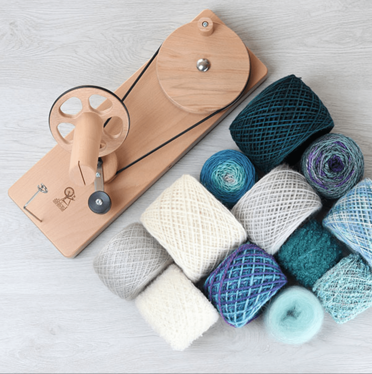 Top-down view of an Ashford Wooden Ball Winder by Ashford Handicrafts Limited, placed beside neatly arranged blue, teal, grey, cream, and white yarn balls on a light wooden surface.
