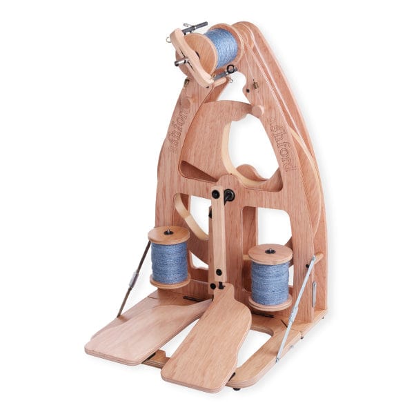 The Ashford Handicrafts Limited Joy 2 Spinning Wheel, with carry bag, features two treadles, three bobbins holding blue-grey fiber, and assorted levers and hooks—ideal for spinning yarn.