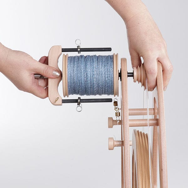 A person adjusts an Ashford Handicrafts Limited Ashford Joy 2 Spinning Wheel with Carry Bag, featuring a bobbin of blue yarn and visible wooden components, demonstrating part of the spinning process.