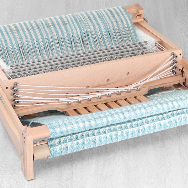 An Ashford Handicrafts Limited Ashford Table Loom, featuring an overhead swinging beater and shuttle race, is set on a light wood surface with blue and white checkered fabric partially woven and rolled on the front beam.