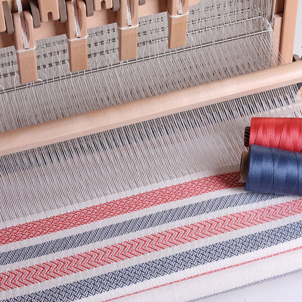 A close-up of an Ashford Handicrafts Limited Table Loom with Overhead Swinging Beater and Shuttle Race, weaving red, blue, and white geometric patterns. Two thread spools—one red, one blue—sit on the right near the woven fabric.