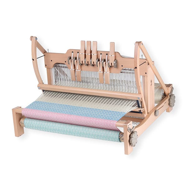 An Ashford Handicrafts Limited Table Loom, with overhead swinging beater and shuttle race, is set on a wooden tabletop, featuring light blue and pink fabric partially woven on the front roller.
