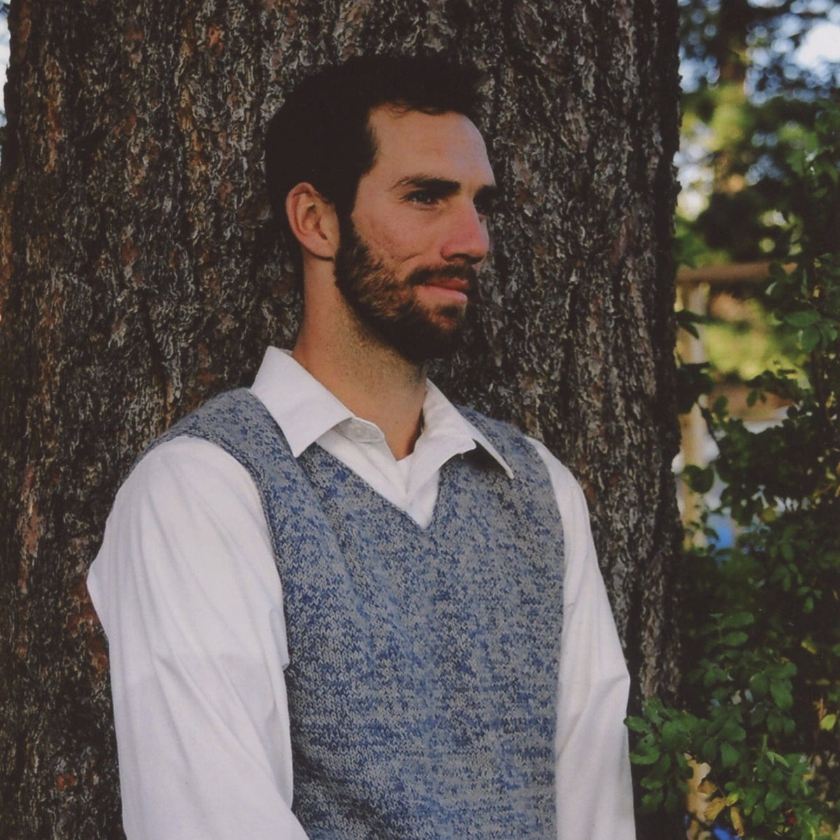 A man with dark hair and a beard, wearing a white shirt and the Bryson Distributing, Inc. Basic Vest for Men with a classic knit pattern, stands outdoors by a tree trunk with green foliage visible nearby.