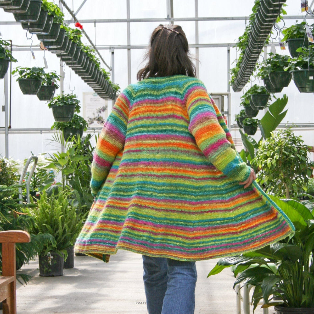A person in blue jeans and the Knitting Pure & Simple Neck Down Swing Coat (#285) walks through a greenhouse filled with hanging and potted green plants. The image, taken from behind, highlights the vibrant cardigan.