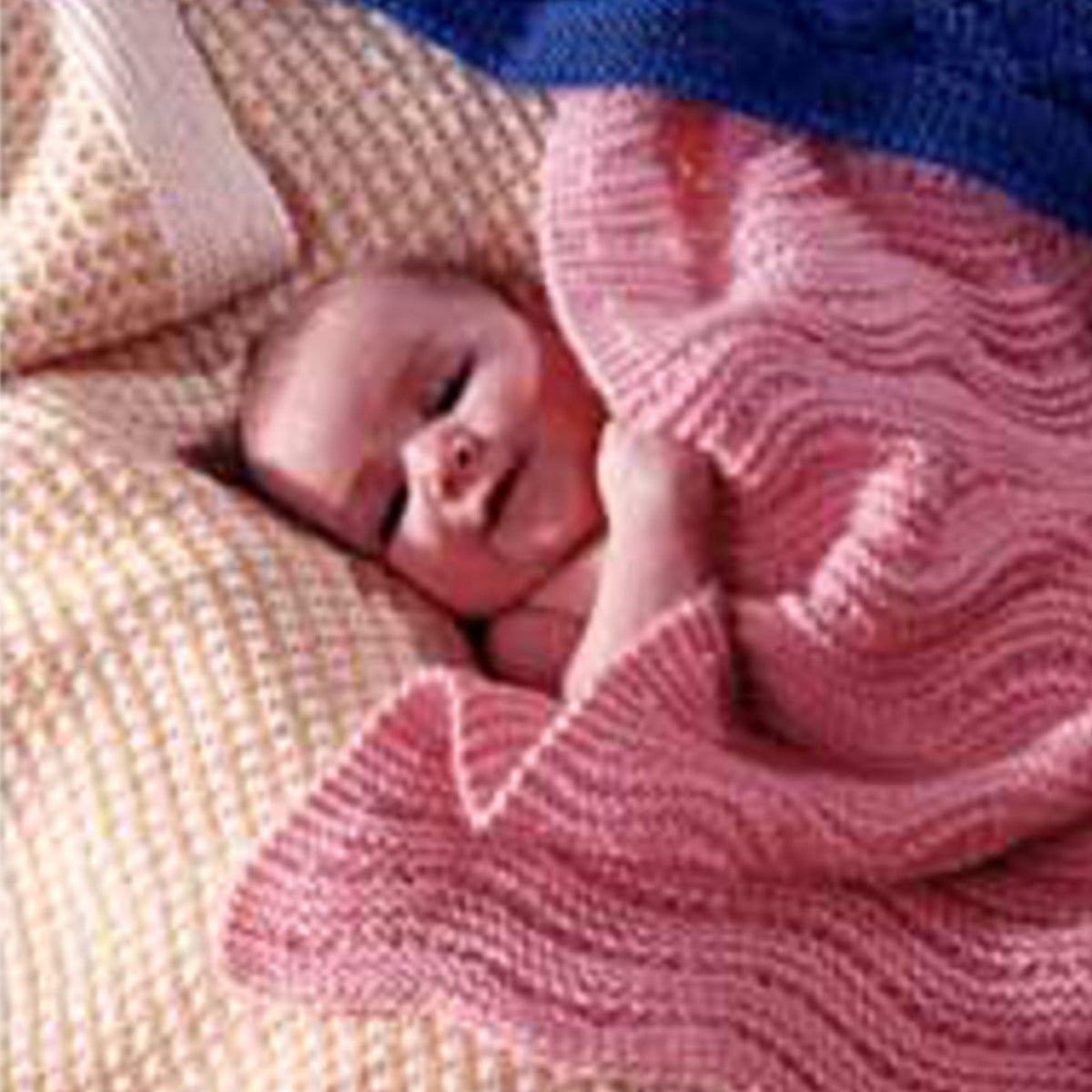 A baby smiles while lying on textured blankets, partially wrapped in the Easy Knit Baby Blankets by Bryson Distributing, Inc. The background includes a cream thermal knit blanket and blue knitted fabric.