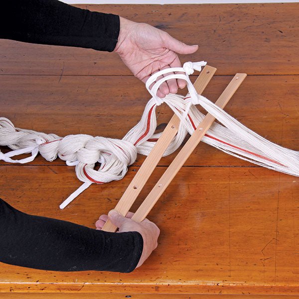 A person threads white and red yarn through holes in Ashford Cross/Warp Sticks for Table Loom by Ashford Handicrafts Limited, preparing a warp on a wooden table.