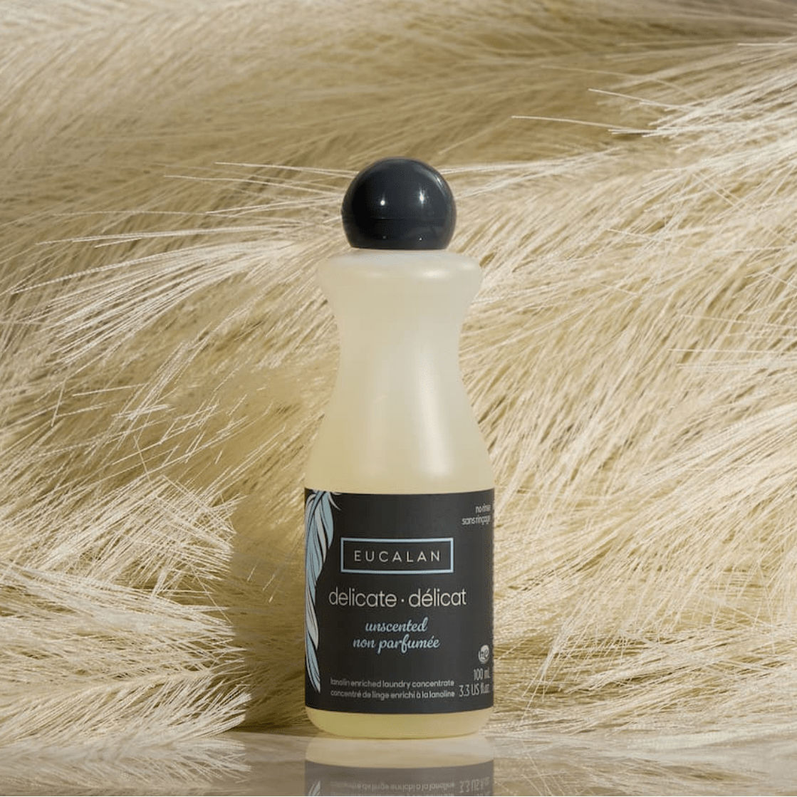 A 100 ml (3.3 oz) bottle of Eucalan Wool Wash by Eucalan is placed on a bed of soft, pale dried grass against a marble-like backdrop.