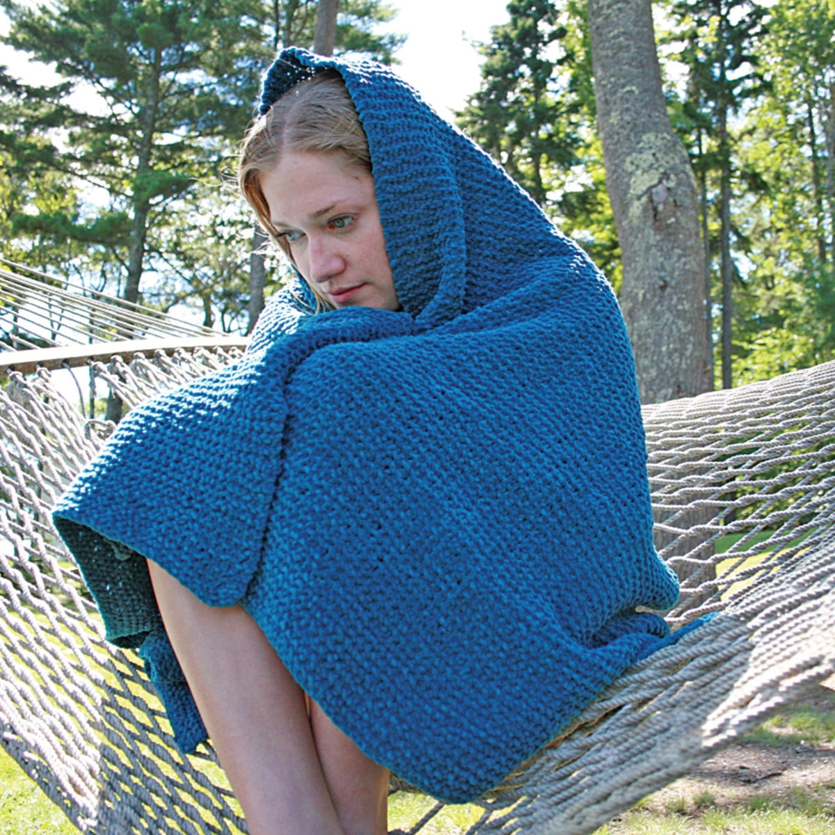 A person sits curled up on a hammock outdoors, wrapped in a Casco Cottage Chenille Throw by Halcyon Yarn, looking thoughtful. Tall trees and greenery can be seen in the background.