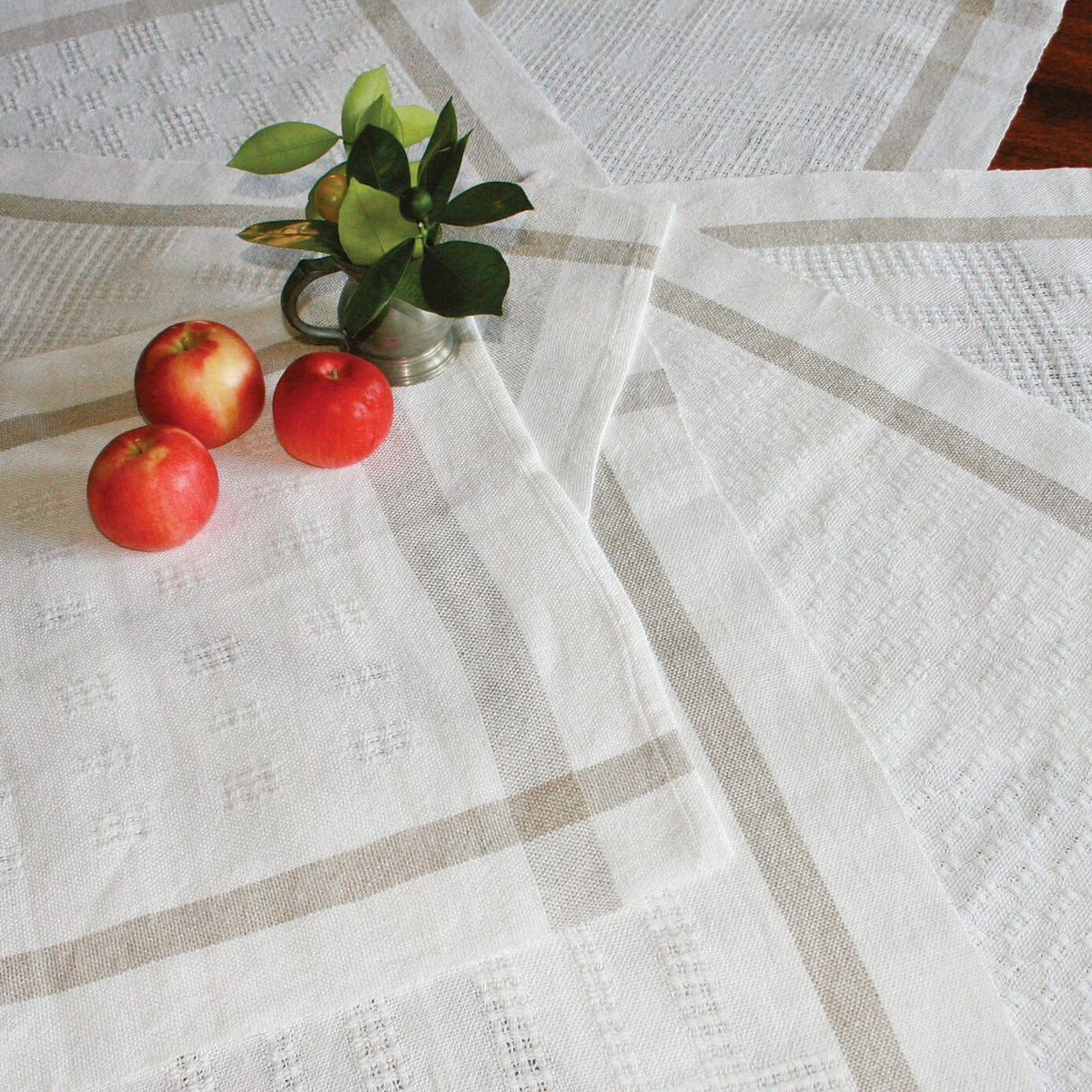 Three red apples and a small vase with green leaves are displayed on Halcyon Yarns Classic Newport Linen Towels, featuring subtle beige stripes and lace patterns, all arranged on a wooden surface.