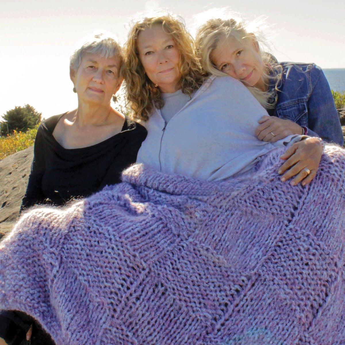 Three women relax outdoors beneath the Halcyon Yarn Cobblestone Cove Victorian Throw, a large textured afghan. Lush greenery and sky set the scene as they enjoy the comfort of this downloadable knitting pattern.