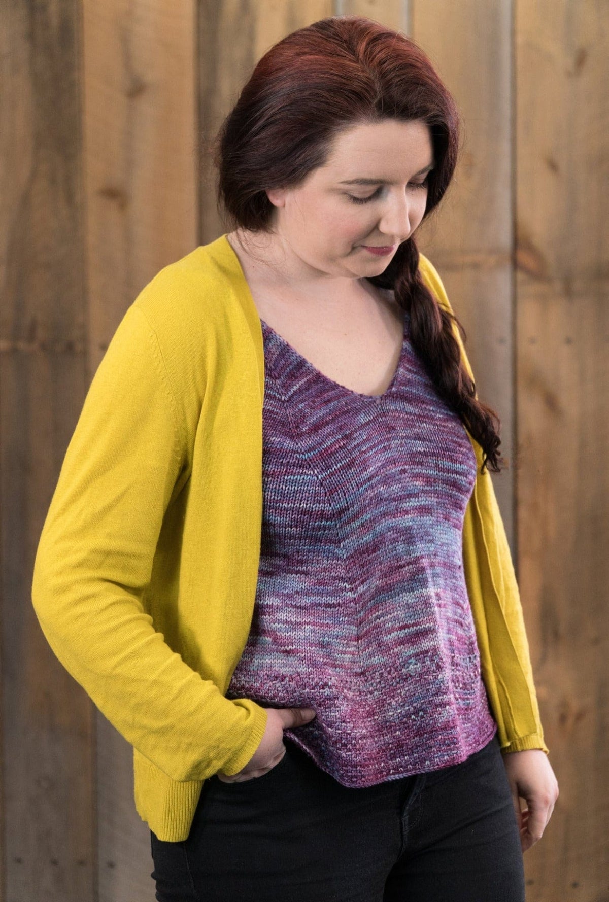 A woman with long brown hair in a braid wears the Flower In The Sun Tank Top by Sierra Roberts, made with Malabrigo Arroyo, layered under a yellow cardigan. She stands against a wooden background, one hand in her pocket, looking down. Halcyon Yarn download.