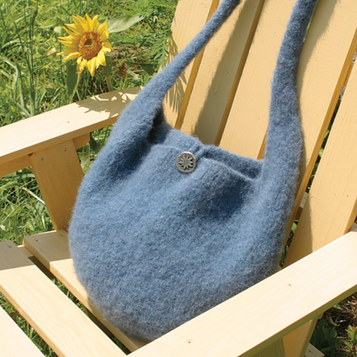 The Halcyon Yarn Felted Satchel Knitted, download, in soft blue bulky wool with a button closure, rests on a light wooden outdoor chair as a sunflower blooms nearby.
