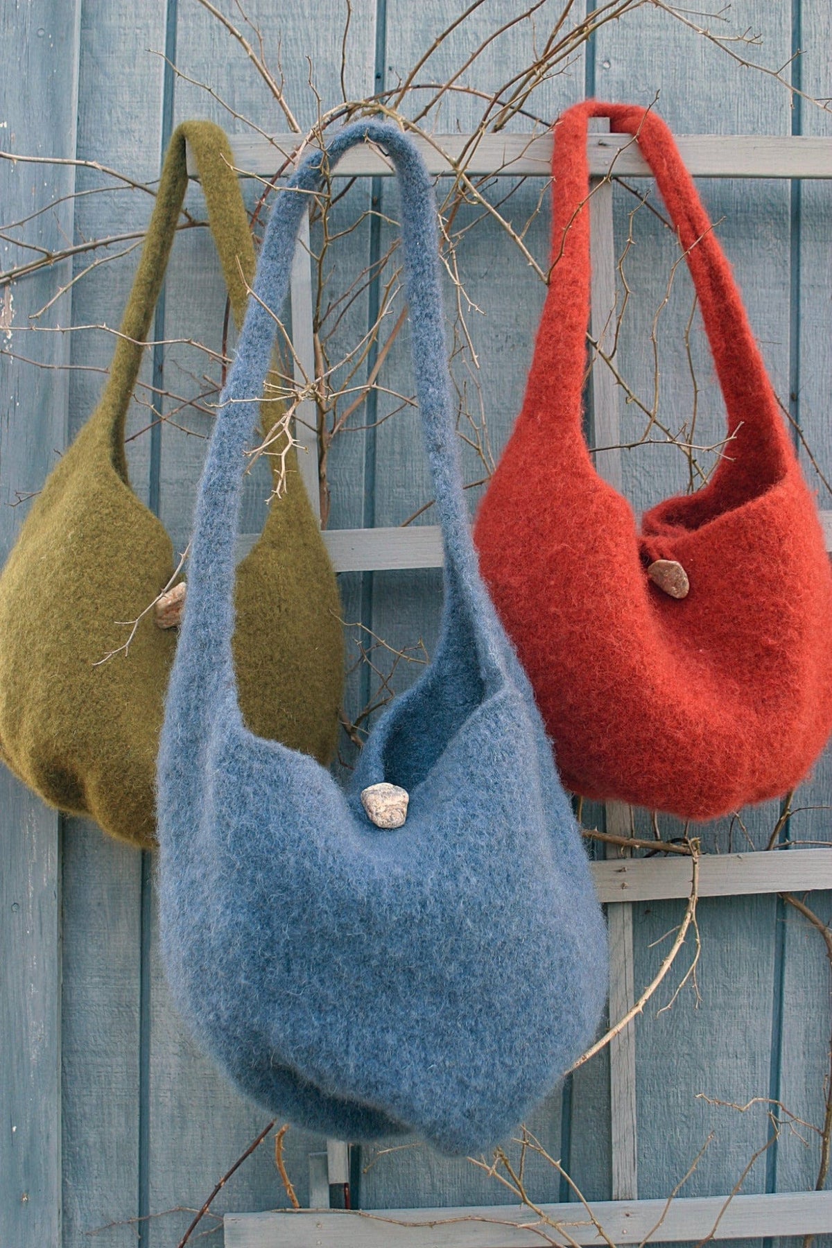 Three Felted Satchel Knitted bags by Halcyon Yarn in olive green, blue, and red hang on a rustic fence. Each wool satchel features a loop closure with a large button—an ideal display of stylish felted knitting. Download available.
