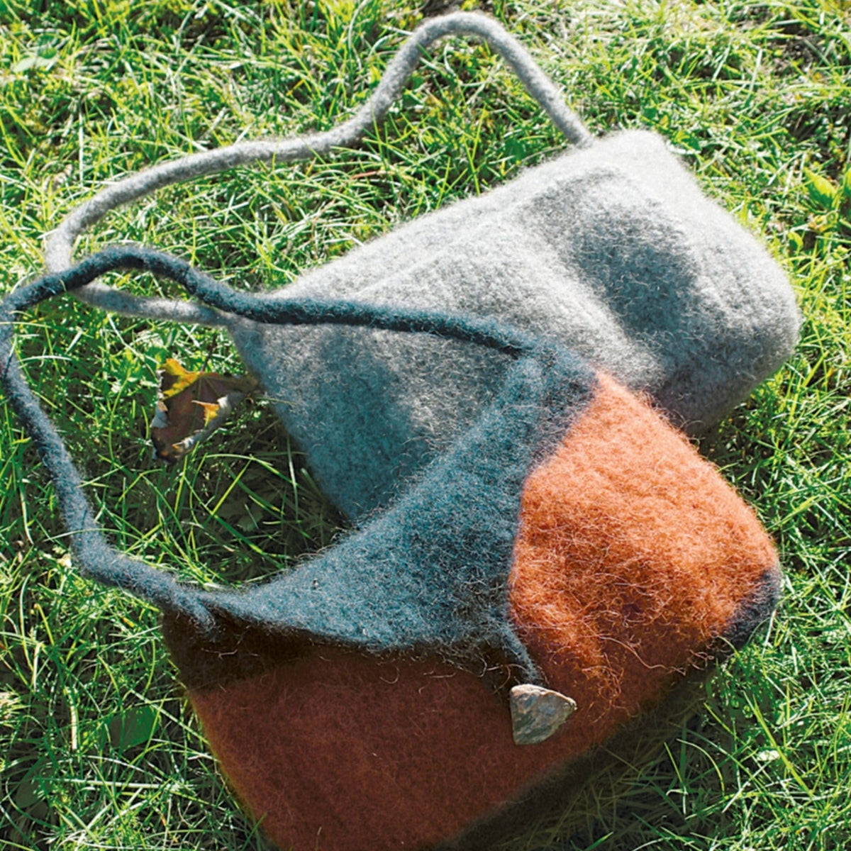 Two Knitted Felt Skinny Strap Shoulder Bags by Halcyon Yarn are on bright green grass: one solid gray, the other color-blocked in rusty orange and gray with a heart-shaped button—both charming examples of felted knitting.