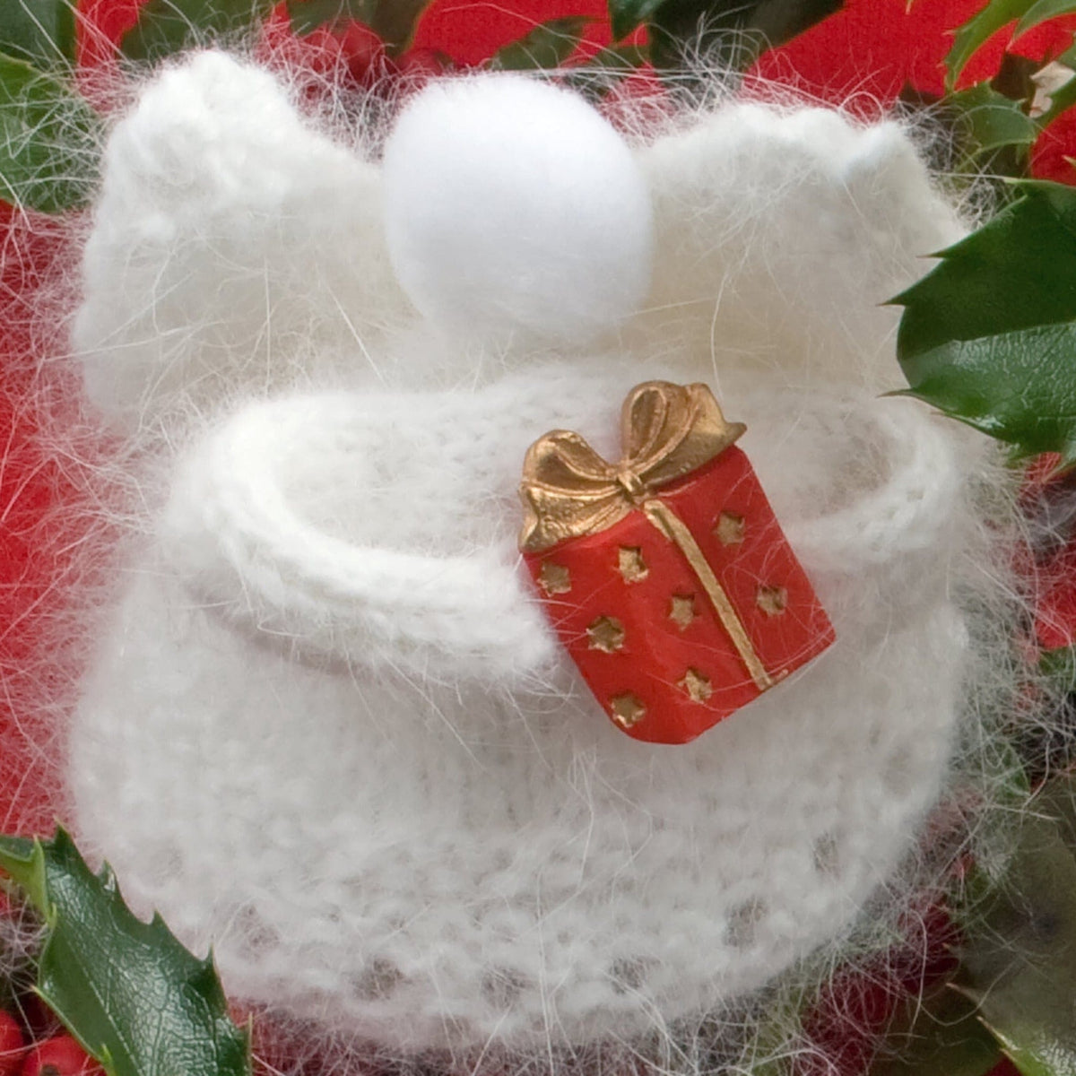 Heavenly Snow Angels by Halcyon Yarn: This small handmade white angora angel with a fluffy pom-pom head holds a red and gold gift box. Surrounded by festive greenery and red accents, it's a charming addition to your holiday ornaments collection. Available for download.