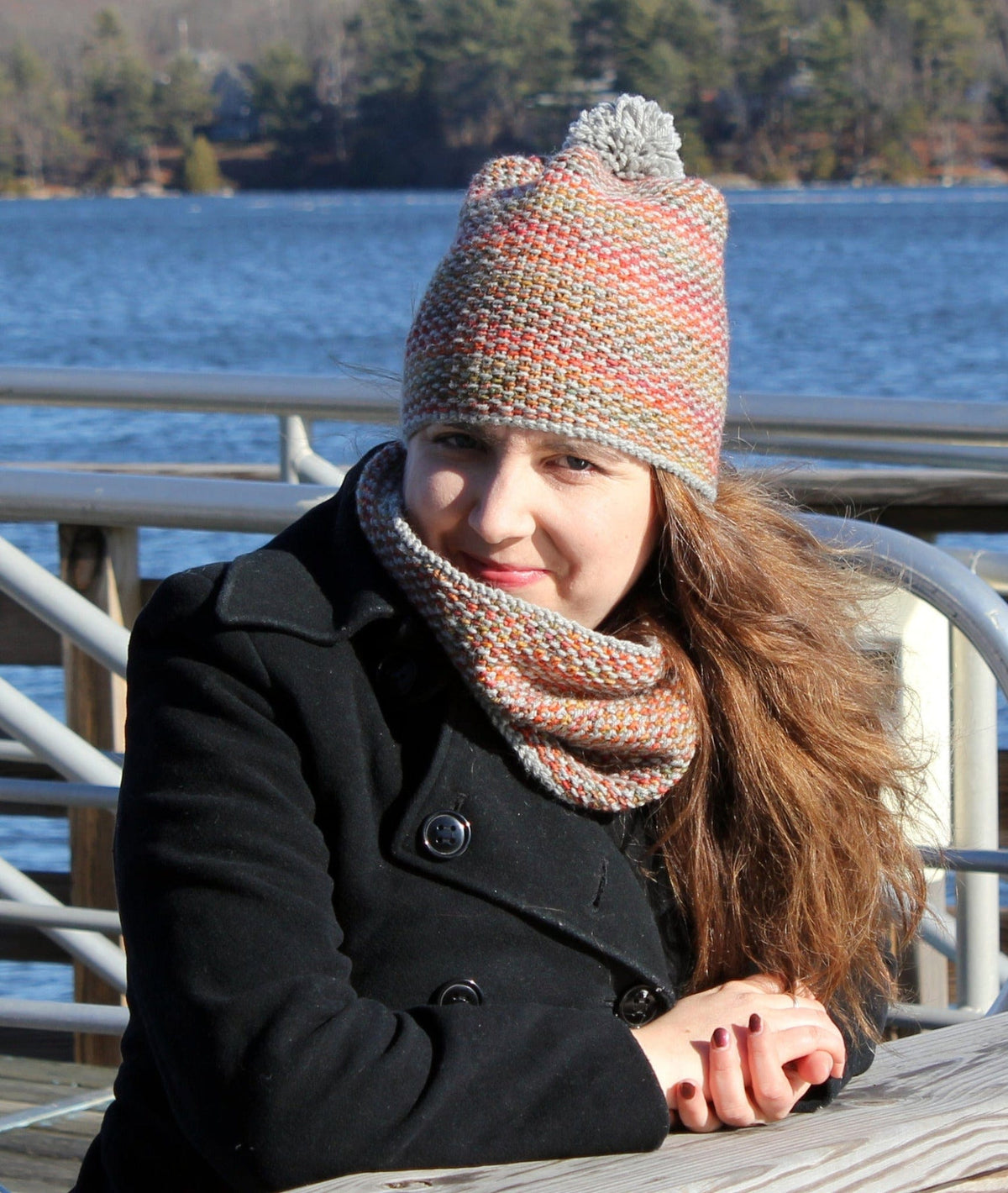 A woman in a black coat wears the Halcyon Yarn Telephone Line Hat & Cowl, download, as she sits at a wooden table by a lake. Her textured knit accessories add warmth and style to the breezy, serene scene.