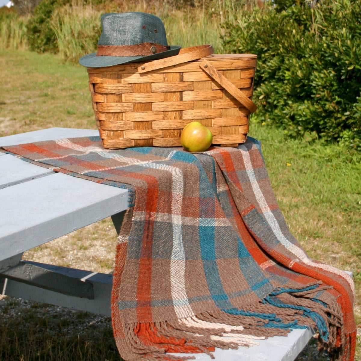 The Fall Picnic Blanket Woven by Halcyon Yarn is spread over a light blue picnic table outdoors, with a basket, green hat, and yellow apple resting on it; grass and bushes create a charming backdrop.