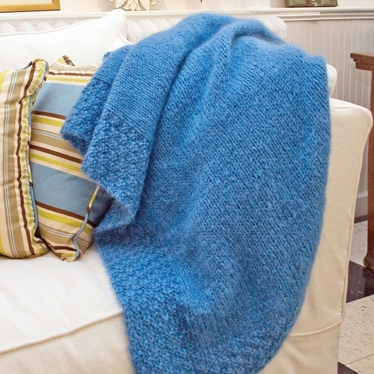 The Halcyon Yarn Victorian Luxury Throw, a textured blue blanket, is draped over the arm of a white couch with striped accent pillows in a cozy living room.
