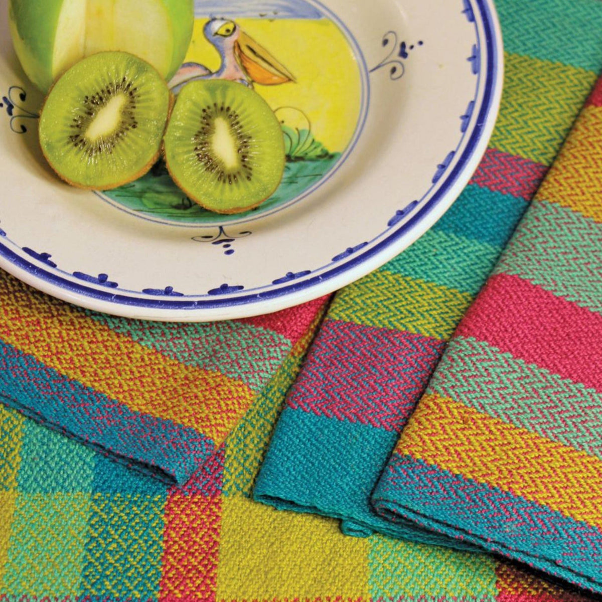 A halved green apple and sliced kiwi rest on a plate with a pelican illustration, atop Halcyon Yarn’s Eight Organic Cottolin Towels featuring colorful, patterned handwoven designs.