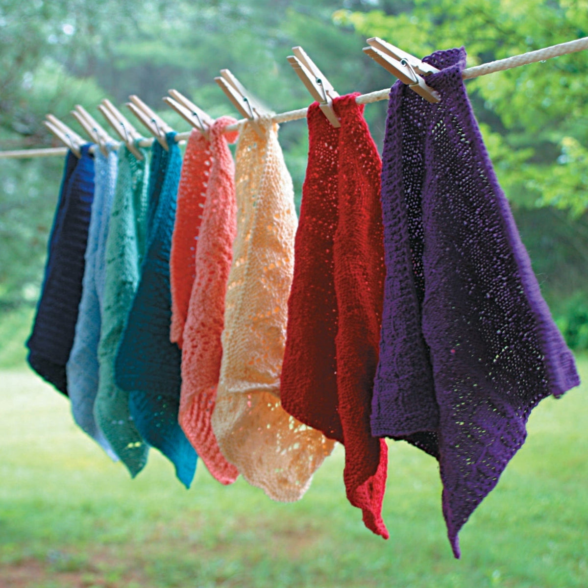 Colorful Casco Bay Washcloths by Halcyon Yarn in blue, green, teal, orange, yellow, red, and purple hang outdoors on a line. These knitted pieces display lovely patterns against a lush green background.