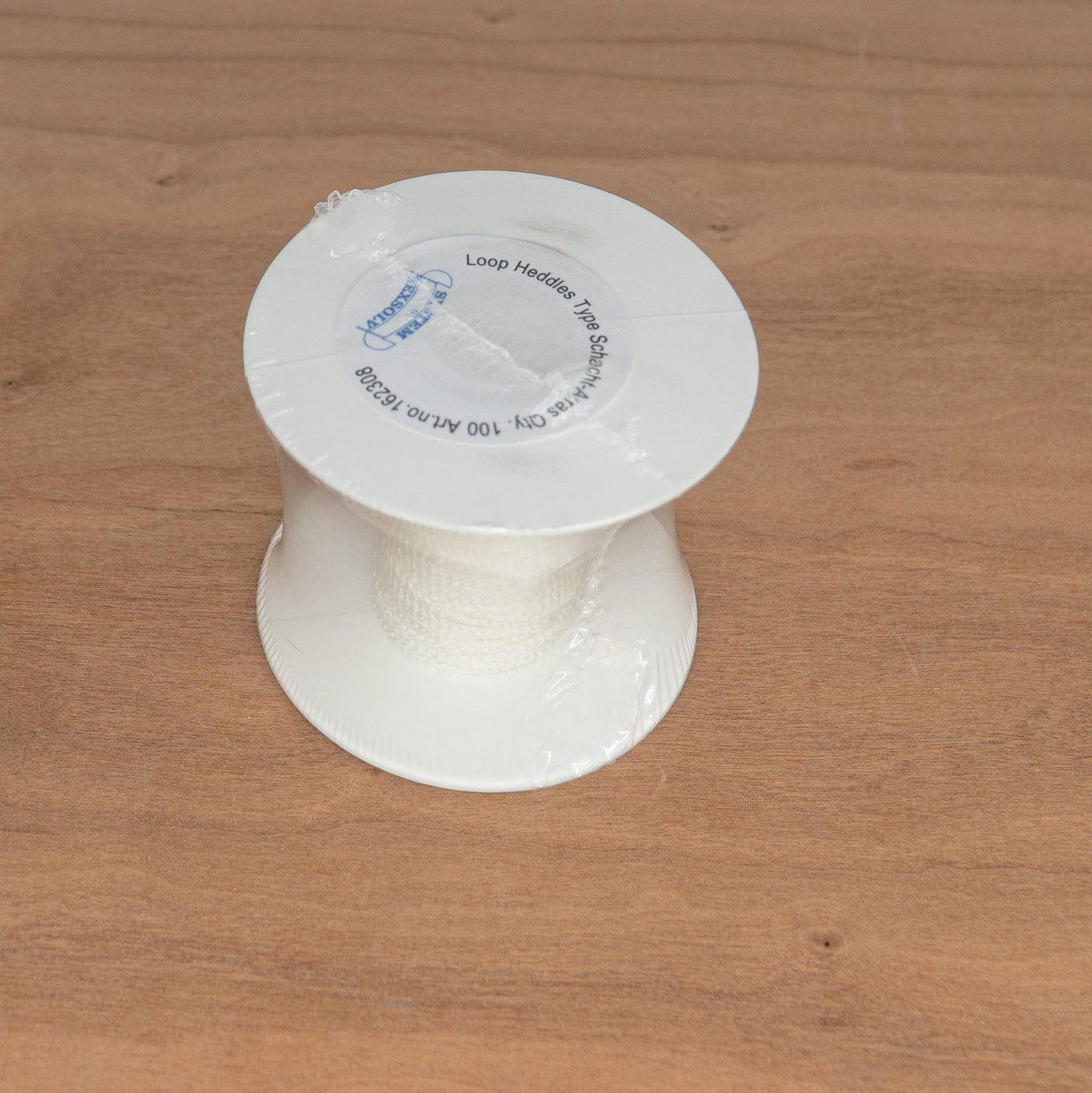 A spool of Texsolv Heddles (4″) by Schacht Spindle Co., designed for use with the Schacht Arras Tapestry Loom, sits partially wrapped in plastic on a light brown wooden surface.