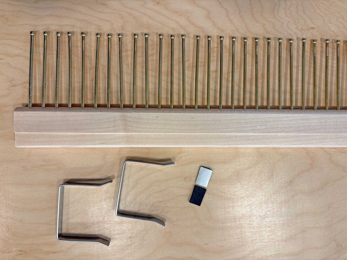 wooden raddle with metal pins set half an inch apart on a wooden background beside small metal clamps.