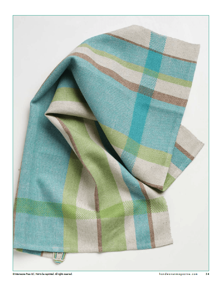 A folded woven fabric in blue, green, beige, and brown plaid on white, evoking Long Thread Media’s "Winning Towels from Handwoven's 21st-Century Towel Contest" (eBook Printed Version) with its soft texture and intersecting lines.