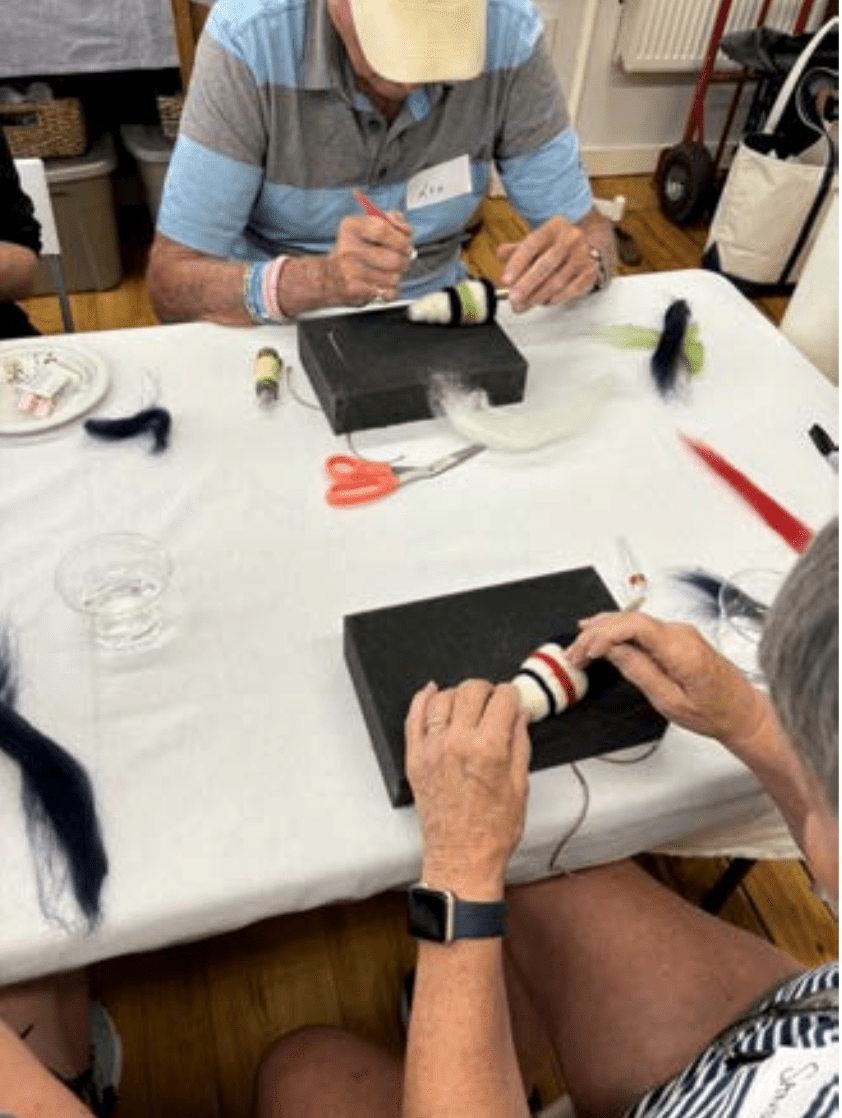 Two people sit at a table in Halcyon Yarn’s "Needle Felting Class: Driftwood Buoys with Michelle Provençal (July 24th)," crafting colorful wool buoys using felting needles, black foam blocks, scissors, and tufts of wool as they focus on their projects.