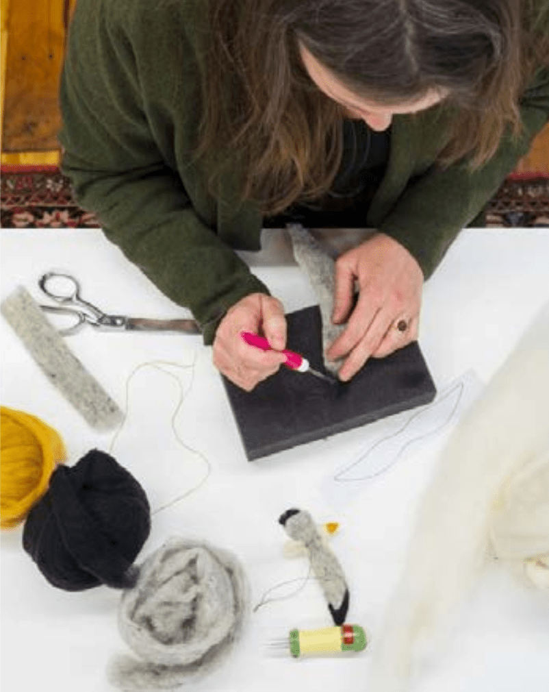 A person needle-felts wool on a pad, surrounded by supplies and a felted bird—ideal for Halcyon Yarn’s "Needle Felting Class: Seagull with Michelle Provençal (July 25th)." Perfect for beginners or seagull ornament making.