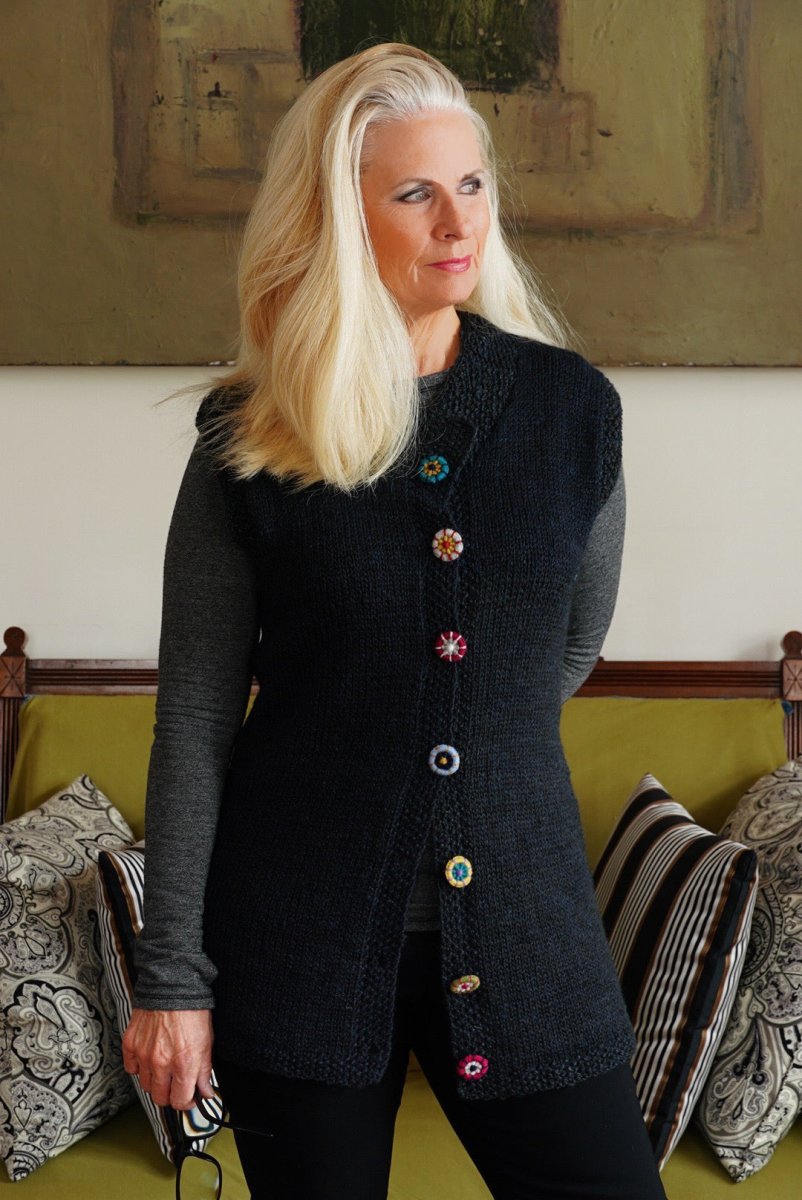 Wearing a sleeveless black sweater decorated with colorful Dorset Buttons, a woman with long blonde hair stands on a sofa with patterned cushions, embodying the creativity found in "Discover Dorset Buttons: A Hands-On Workshop with Linda Perry (October 24, 2026)" by Halcyon Yarn.