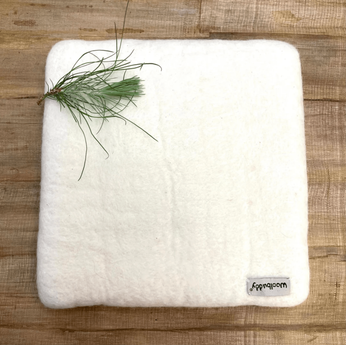 The Woolbuddy Felting Mat, Small, is a square white mat made from 100% wool by Woolbuddy, featuring a small fabric tag and shown on wood with a green pine twig in the corner.