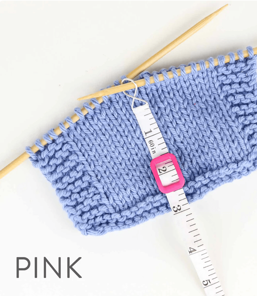 A swatch of light purple knitting on wooden needles features the Twice Sheared Sheep Work Until... Measuring Tape (white) and a pink stitch marker showing 2 inches. The word PINK appears in gray text in the corner.