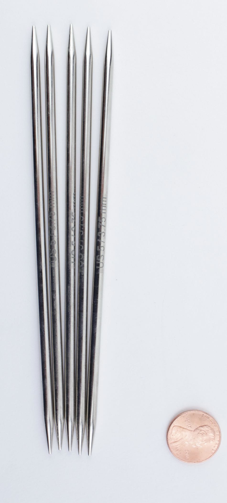 Five Knit Pro Nova Metal Double-Point Knitting Needles (6 inch, Accessories Unlimited) are arranged side by side on a white background, with a penny at the lower right for scale.