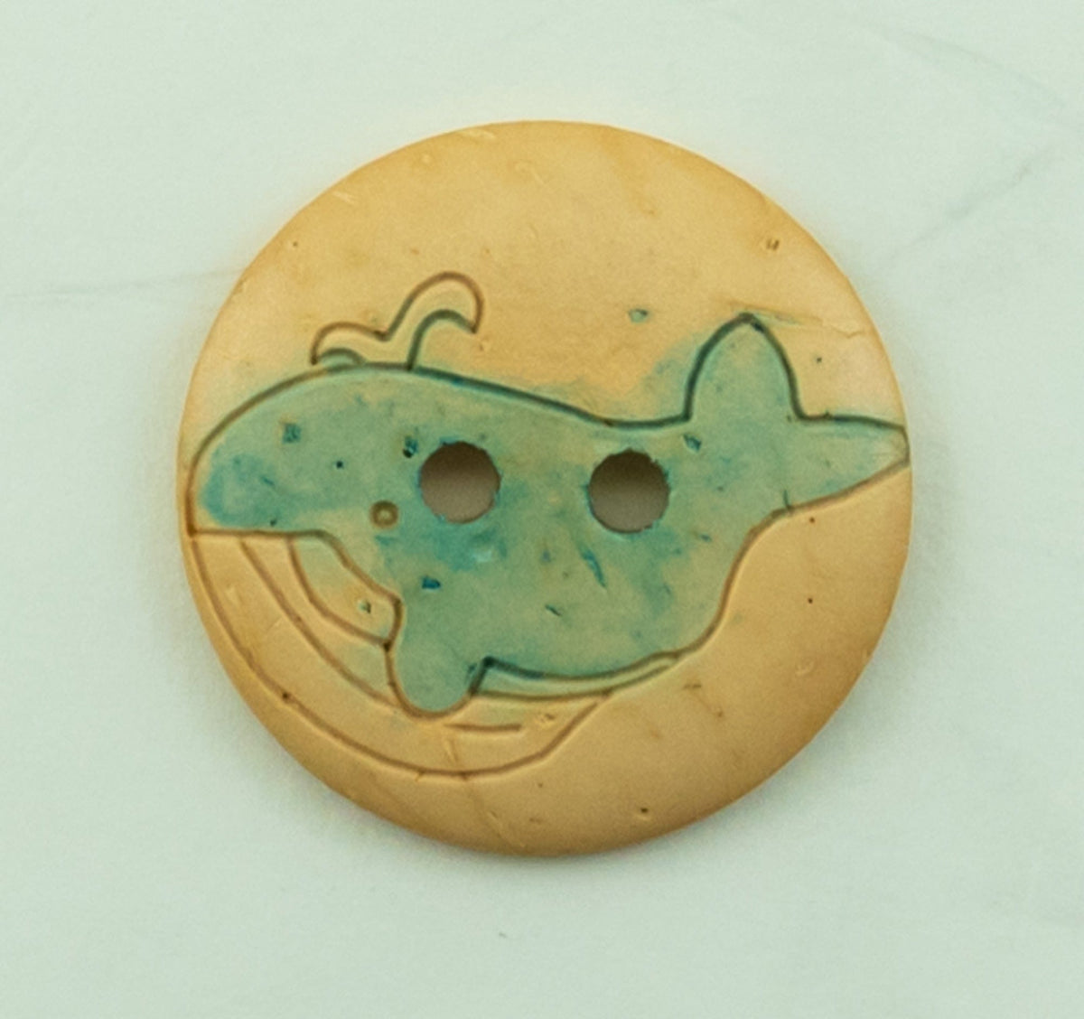 The Buttons Etc. Baby Animal Button, 13/16", is a round beige sewing button with two holes, featuring an engraved and painted blue whale with a water spout and a rainbow pattern in the background.