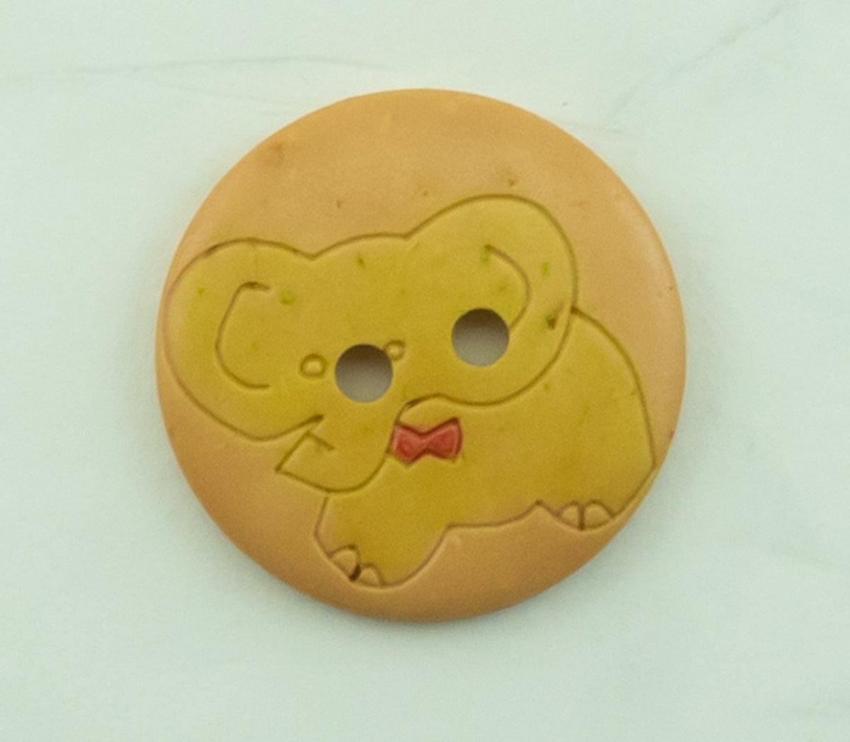 The Buttons Etc. Baby Animal Button, 13/16", is a round light brown button with two holes and an engraved elephant wearing a red bow tie, shown on a light surface.