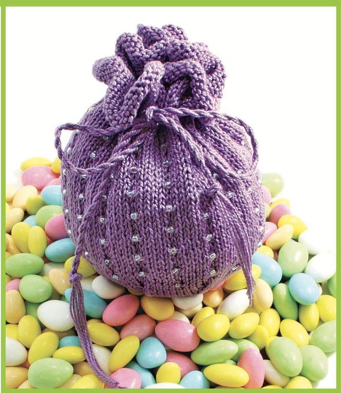 A vibrant purple Cotton Jelly Bean Bag, crafted with a drawstring closure from Halcyon Yarn, rests atop a pile of colorful candy-coated almonds, set against a white background framed with a green border.