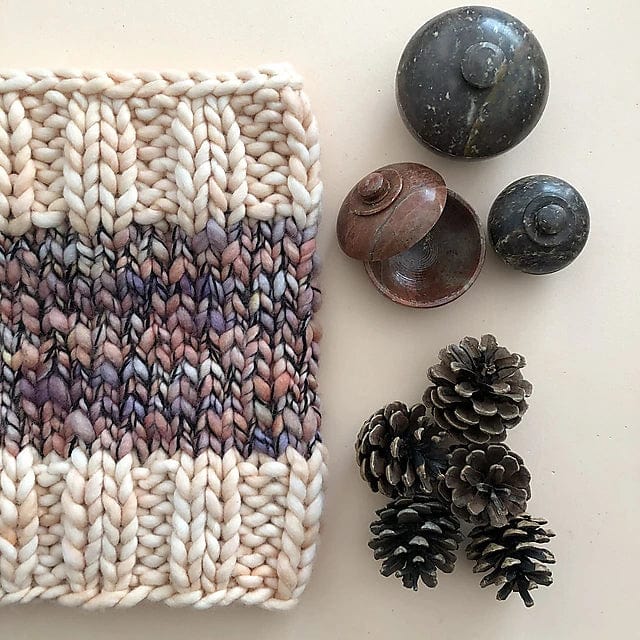 The Aguas Blancas Cowl Pattern by Kristel Nieves for Ravelry Instore features chunky knitted fabric in cream and purple-brown merino wool, displayed beside pinecones and dark containers—perfect for a cozy knit project.