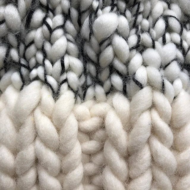 Close-up of chunky knitted fabric made with Malabrigo yarn, showing white-black mixed fibers on top and solid cream below. The cozy merino texture suggests it’s crafted using the Aguas Blancas Hat Pattern by Kristel Nieves from Ravelry Instore.
