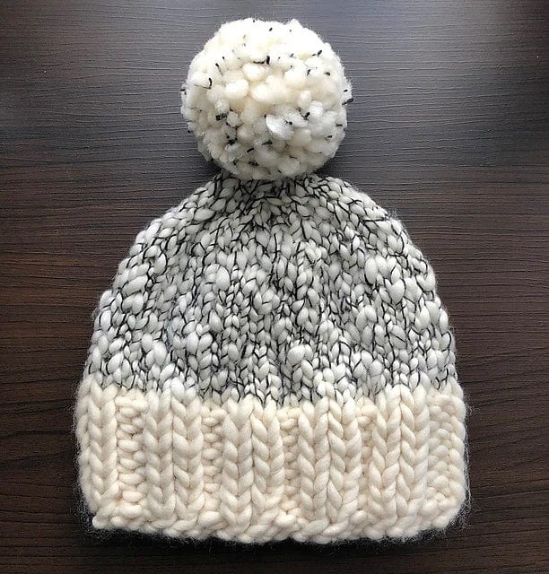 An Aguas Blancas Hat made from the pattern by Kristel Nieves (Ravelry Instore), featuring chunky white and gray yarn, a ribbed brim, and a large pom-pom, displayed on a dark wooden surface.