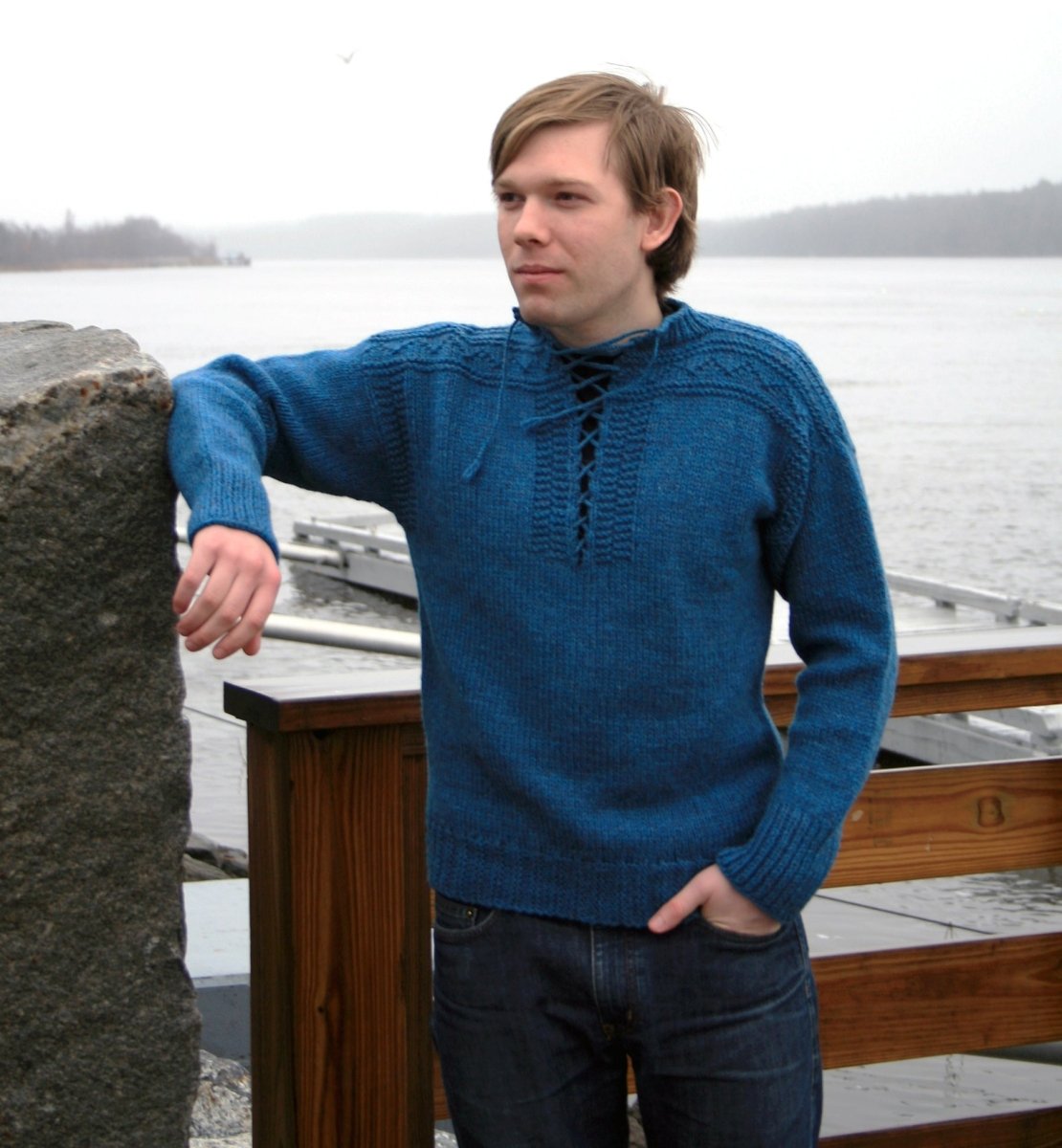 Wearing the Halcyon Yarn Portuguese Fisherman Pullover, a person stands by a wooden fence near a body of water, resting one arm on a stone post. Trees and docks fade into the misty background, creating a serene setting ideal for family outings or beginner-friendly adventures. Download available.