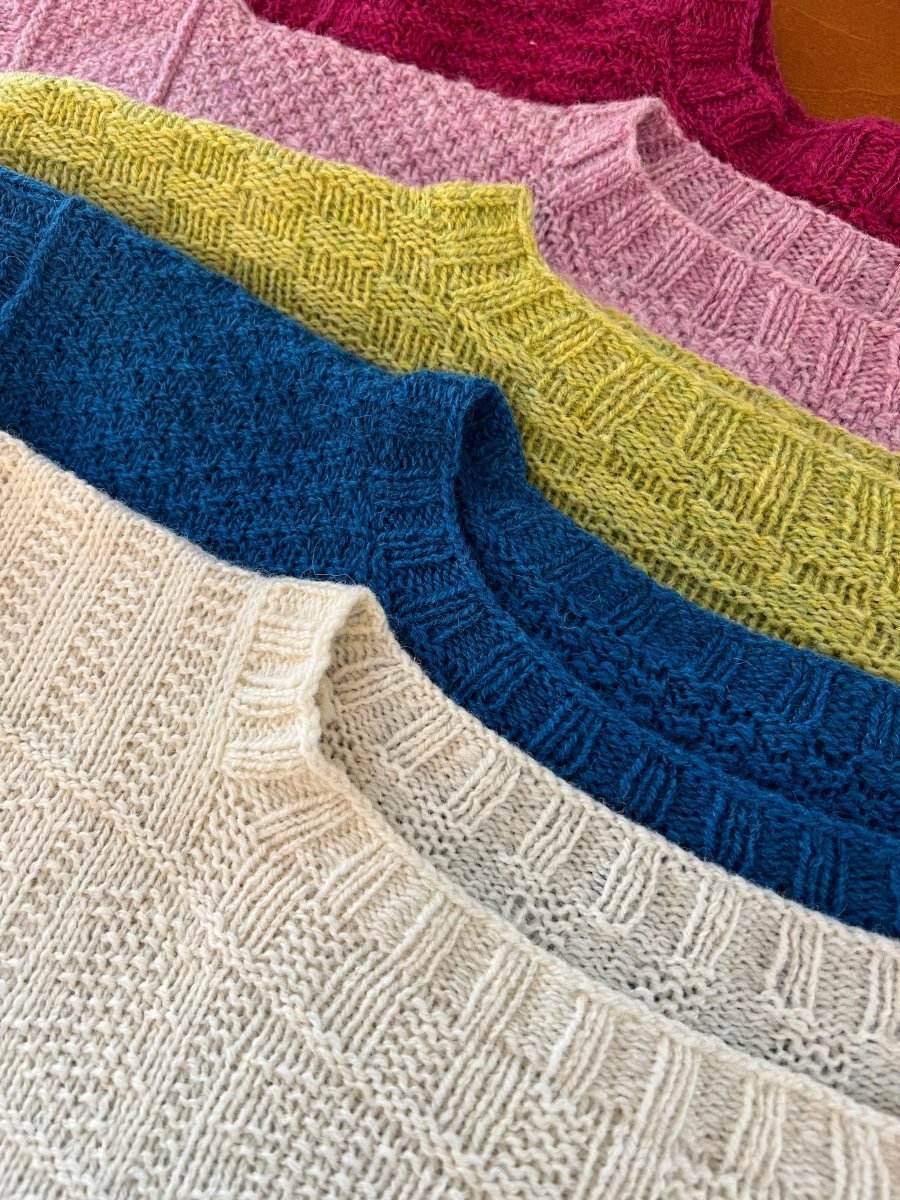 Five hand-dyed Gansey sweaters in cream, blue, yellow, light pink, and magenta—featuring textured knit patterns—are stacked for Halcyon Yarn's Pop-up Sale with Linda Perry on Saturday, May 23rd.