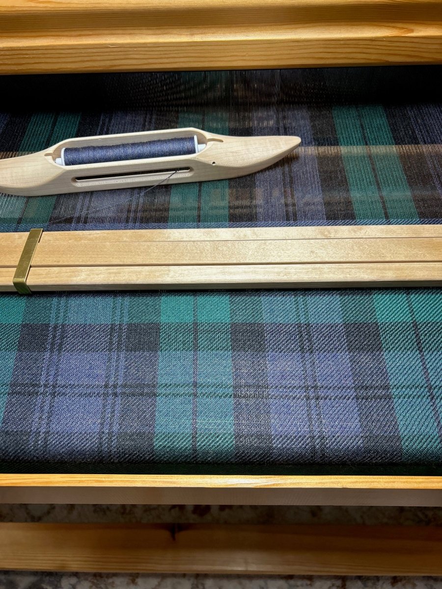At Halcyon Yarn’s Weaving Demo + Pop-Up Shop for Oasis Free Clinic (May 8-9), a wooden shuttle and reed rest on a loom as vibrant blue, green, and black tartan plaid fabric is woven live.