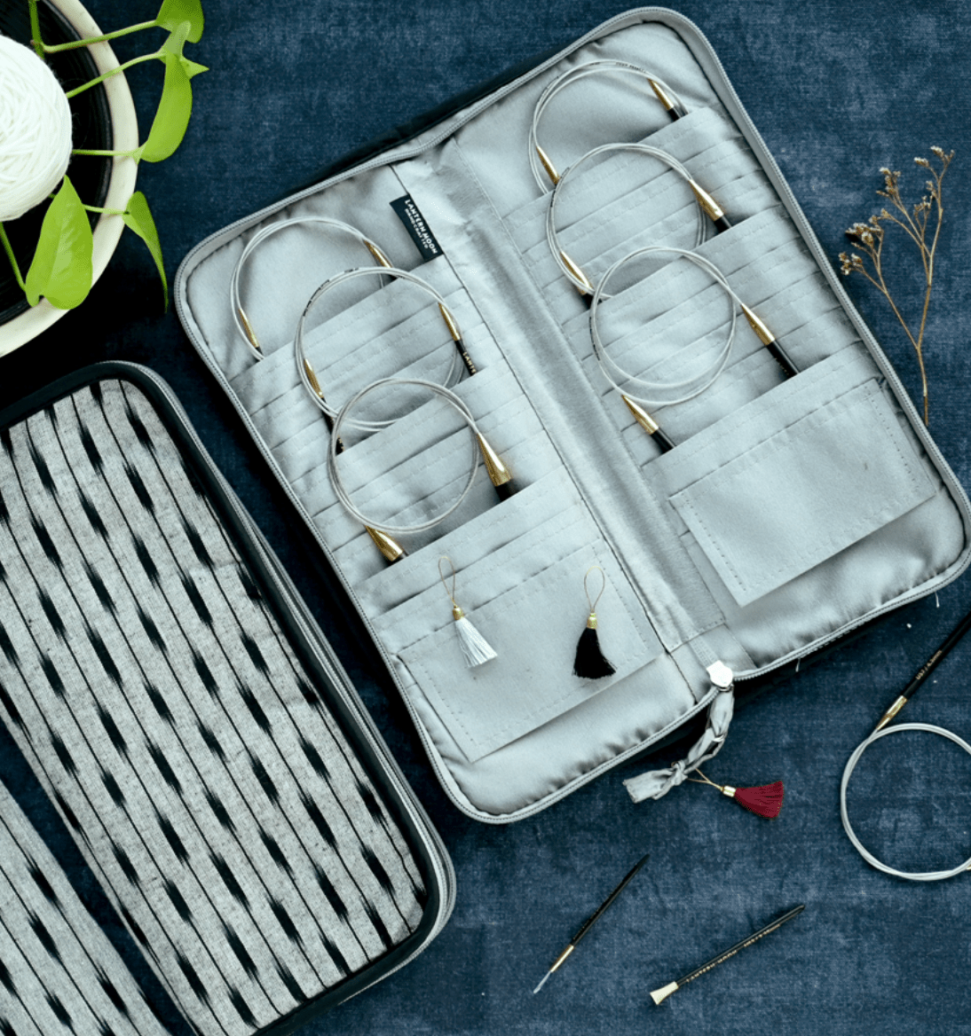 The Lantern Moon Circular Needle Case by Bryson Distributing, Inc. stores your circular knitting needles and stitch markers in style, resting open on handmade IKAT fabric beside thick yarn, a white skein, a plant, and dried flowers.