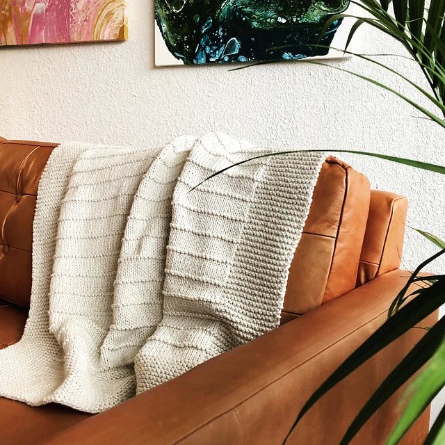 The On The Porch Baby Blanket Pattern by Fifty Four Ten Studio (print) from Ravelry Instore is draped over a tan leather couch, with a green plant in front and colorful abstract art on the white wall behind.