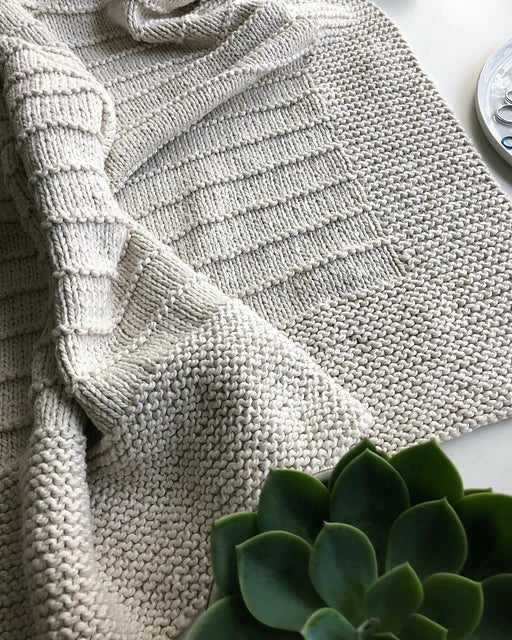 A close-up of the On The Porch Baby Blanket Pattern by Fifty Four Ten Studio (print) from Ravelry Instore, showing its textured beige knit partially folded on white, with a green succulent and thread container nearby.